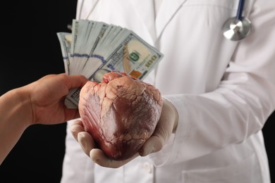 Photo of Patient giving dollar banknotes to doctor with heart on black background, closeup