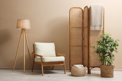 Photo of Folding screen, blanket, armchair, lamp and green houseplant near beige wall indoors