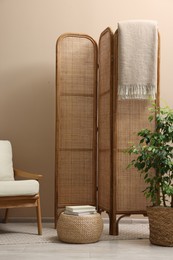 Folding screen, blanket, armchair and green houseplant near beige wall indoors