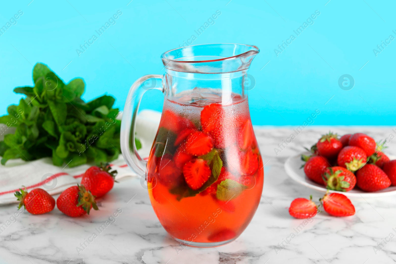 Photo of Tasty strawberry lemonade with mint in jug and berries on white marble table against light blue background