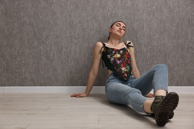 Photo of Beautiful young woman in stylish corset on floor near grey wall. Space for text
