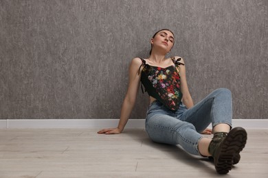 Beautiful young woman in stylish corset on floor near grey wall. Space for text