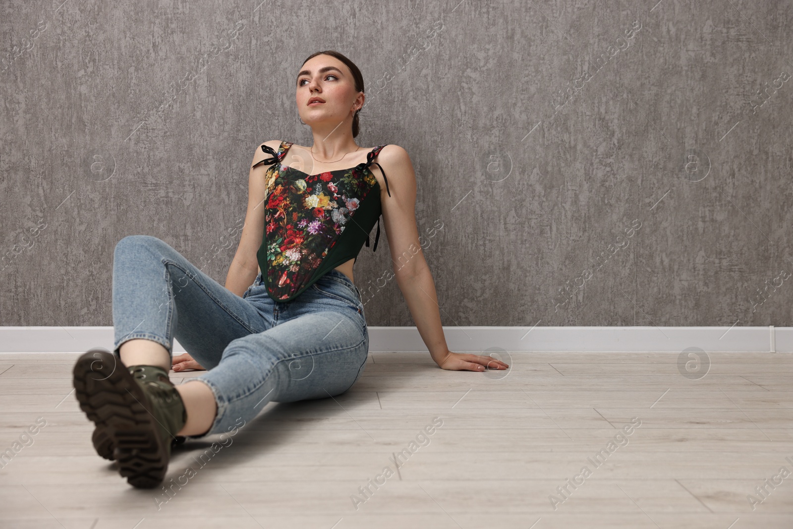 Photo of Beautiful young woman in stylish corset on floor near grey wall. Space for text