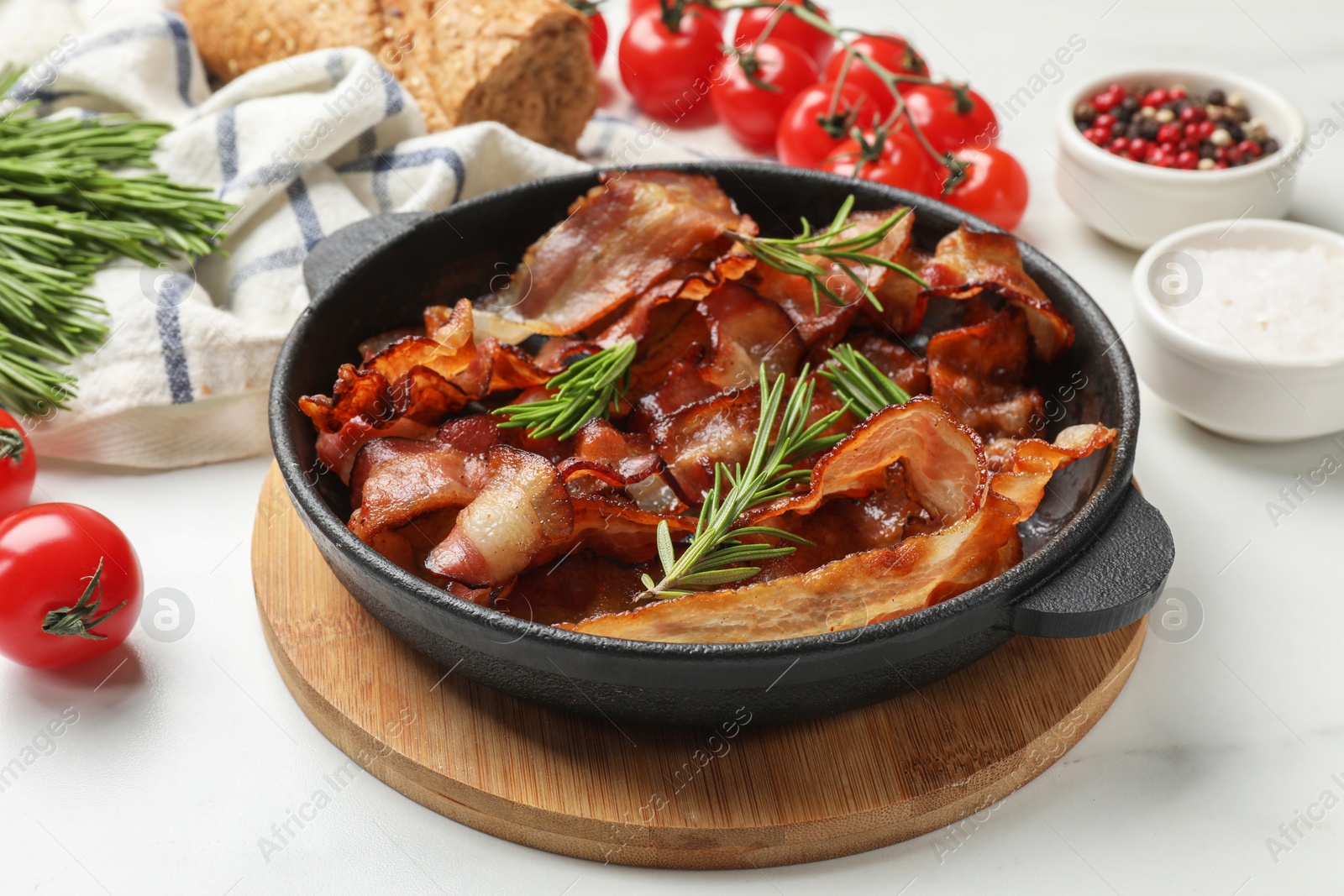 Photo of Delicious bacon slices in frying pan, spices and tomatoes on white table