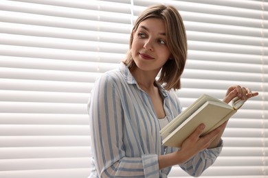 Photo of Woman reading book near window blinds at home, space for text