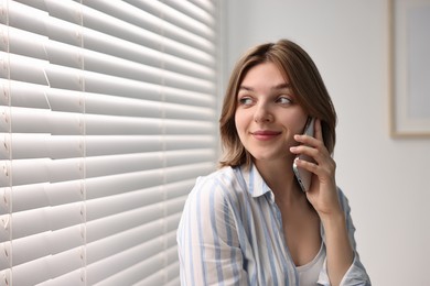 Woman talking on phone near window blinds at home, space for text