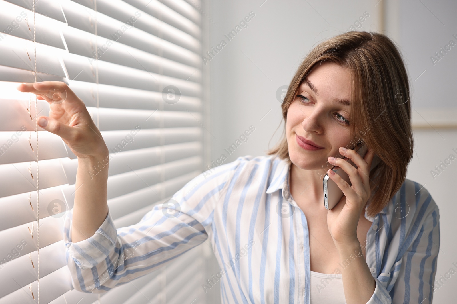 Photo of Woman talking on phone near window blinds at home