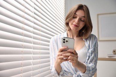 Woman using phone near window blinds at home, space for text