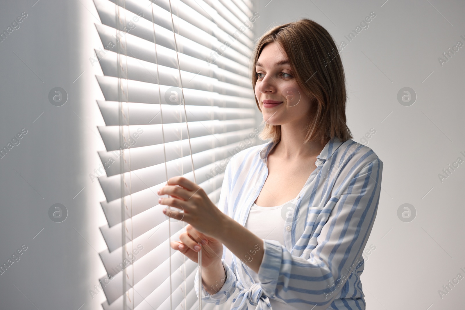 Photo of Young woman adjusting window blinds at home