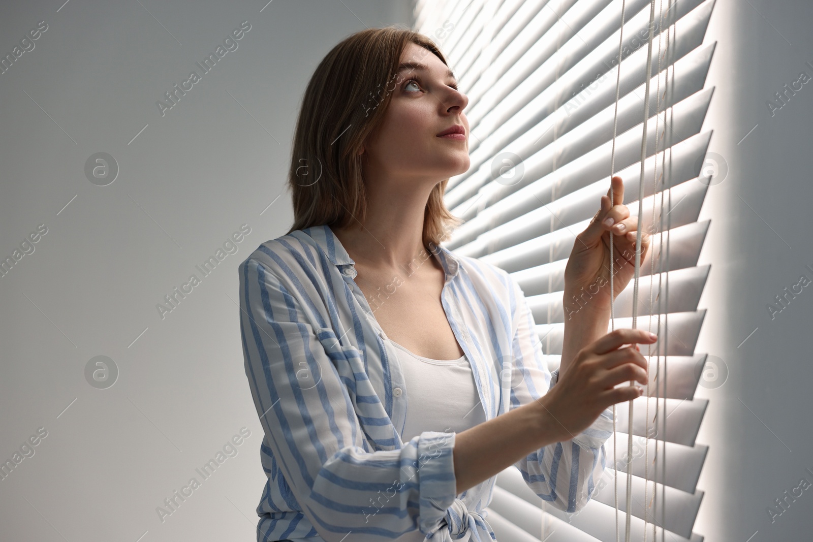 Photo of Young woman adjusting window blinds at home