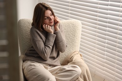 Woman sitting on armchair near window blinds at home, space for text