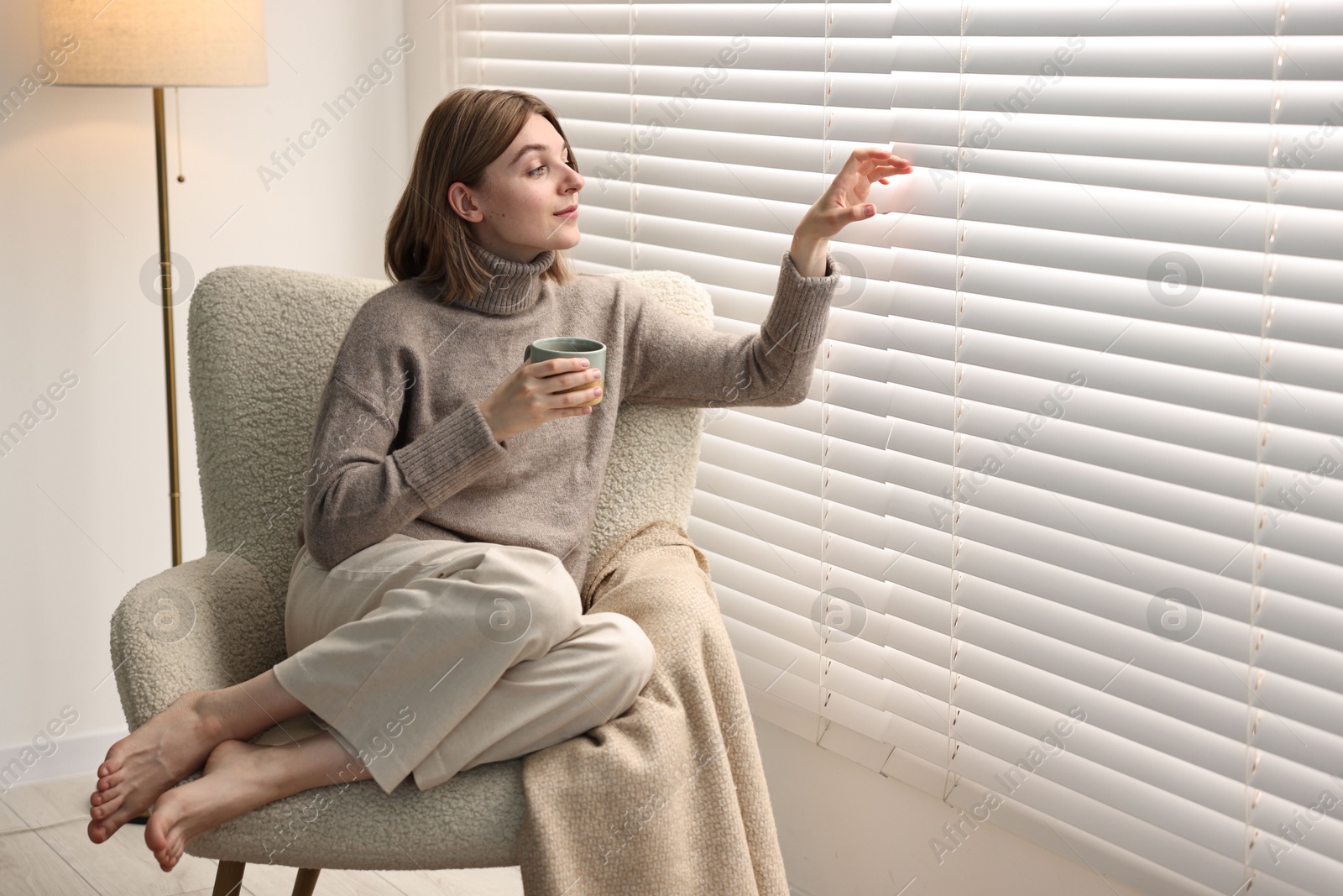 Photo of Woman with cup of drink sitting on armchair near window blinds indoors