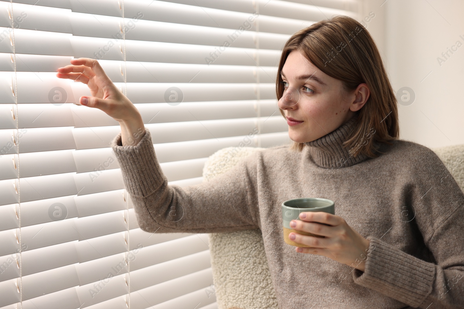 Photo of Woman with cup of drink sitting on armchair near window blinds indoors