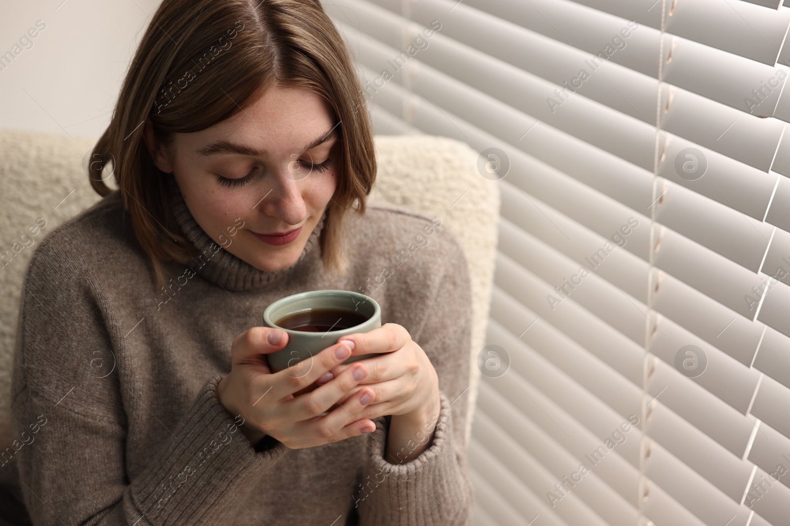 Photo of Woman with cup of drink sitting on armchair near window blinds indoors