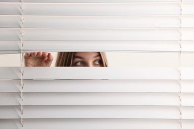 Young woman looking through window blinds on white background, space for text