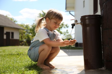 Photo of Water scarcity. Cute little girl drawing water with hands from tap outdoors