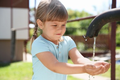 Photo of Water scarcity. Cute little girl drawing water with hands from tap outdoors