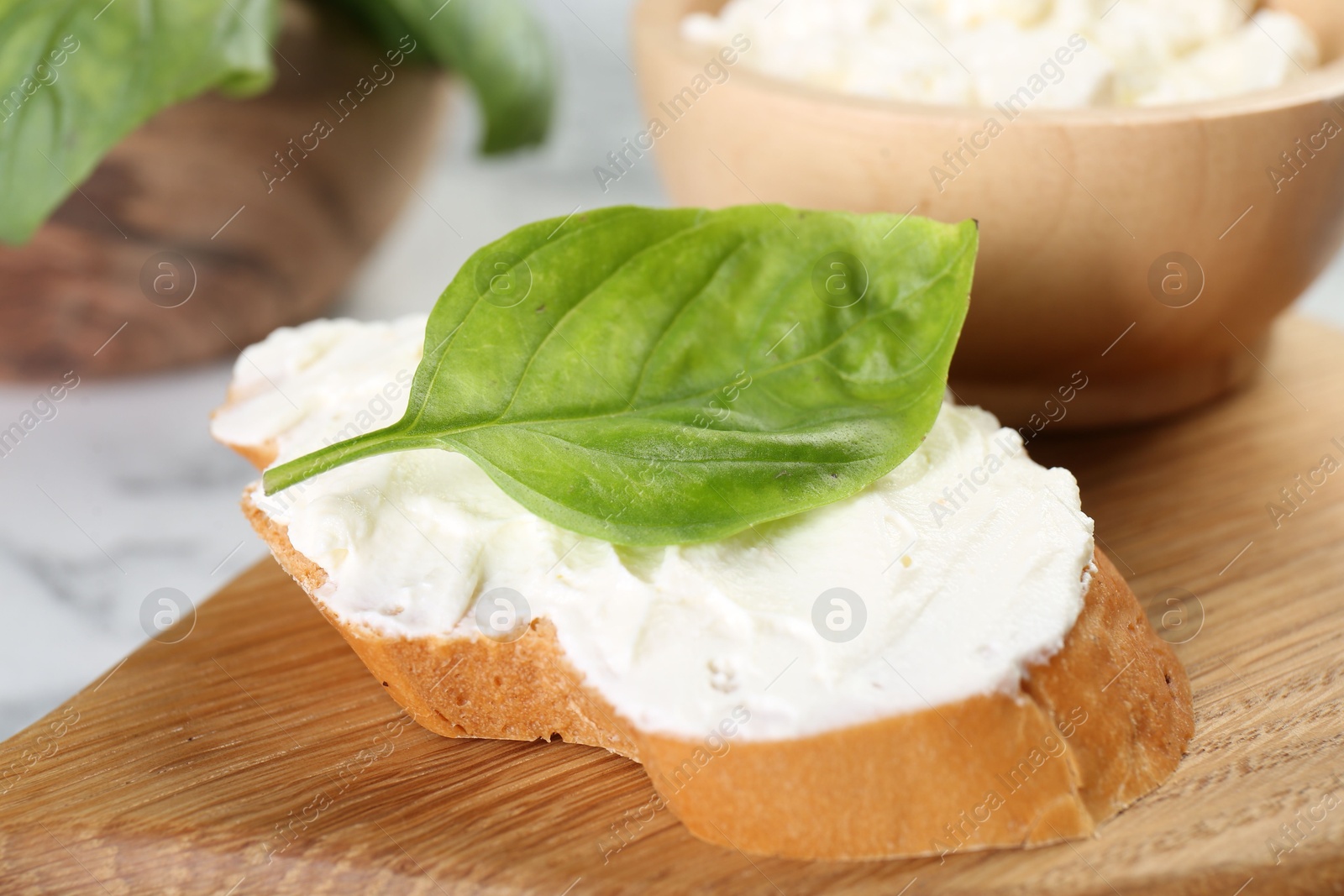 Photo of Piece of bread with cream cheese and basil leaf on white table, closeup