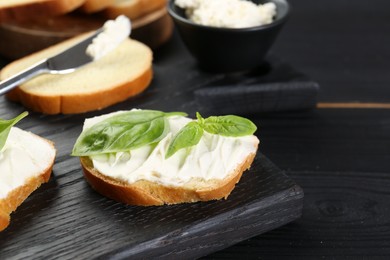 Pieces of bread with cream cheese and basil on black wooden table. Space for text