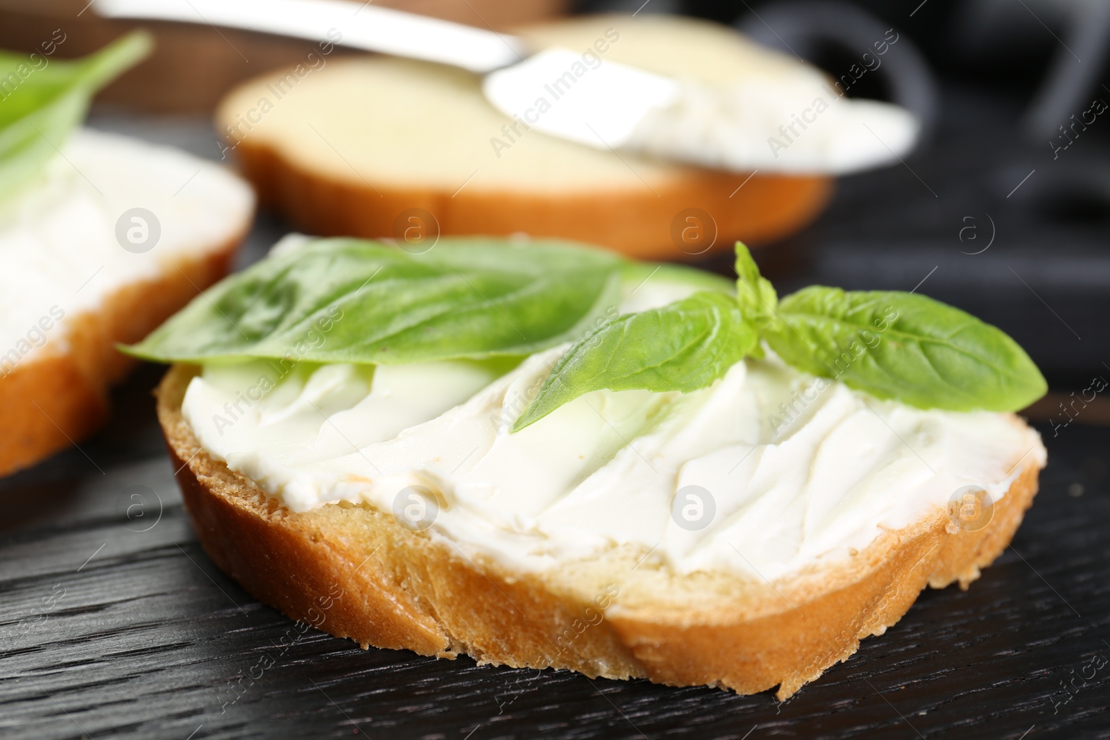 Photo of Piece of bread with cream cheese and basil on black wooden table, closeup