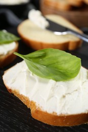 Piece of bread with cream cheese and basil on black table, closeup