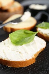 Piece of bread with cream cheese and basil on black table, closeup