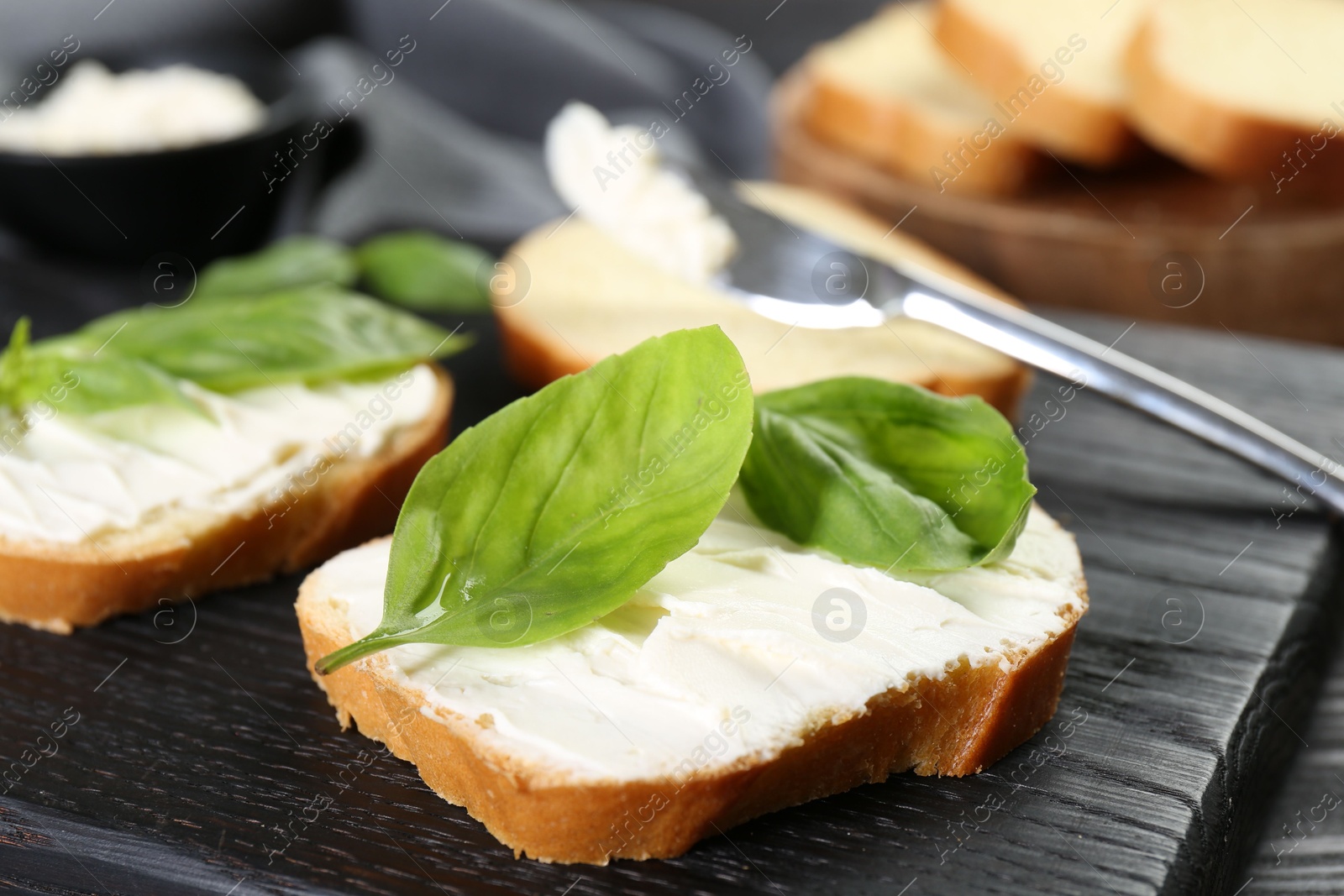 Photo of Pieces of bread with cream cheese and basil leaves on black table, closeup