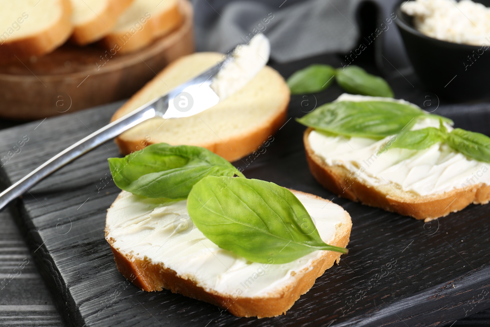 Photo of Pieces of bread with cream cheese and basil leaves on black table, closeup