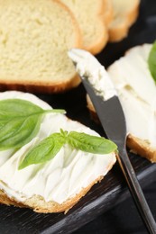 Photo of Pieces of bread with cream cheese, knife and basil leaves on black table, closeup