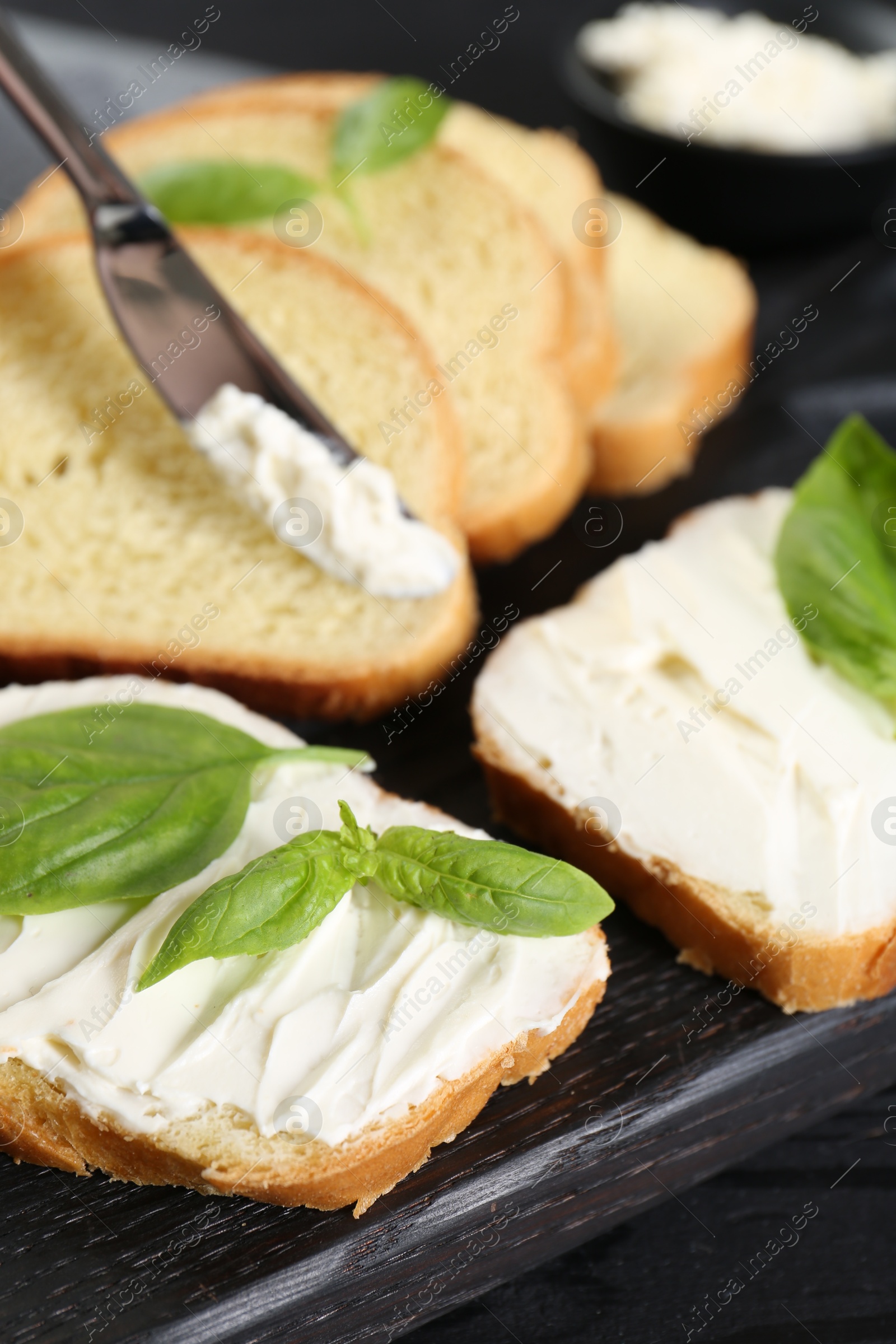 Photo of Pieces of bread with cream cheese and basil leaves on black table, closeup