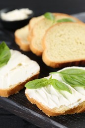 Photo of Pieces of bread with cream cheese and basil leaves on black table, closeup