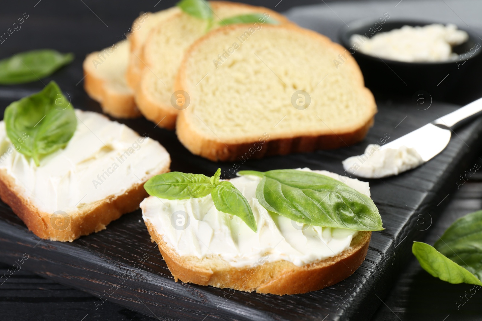 Photo of Pieces of bread with cream cheese and basil leaves on black table, closeup