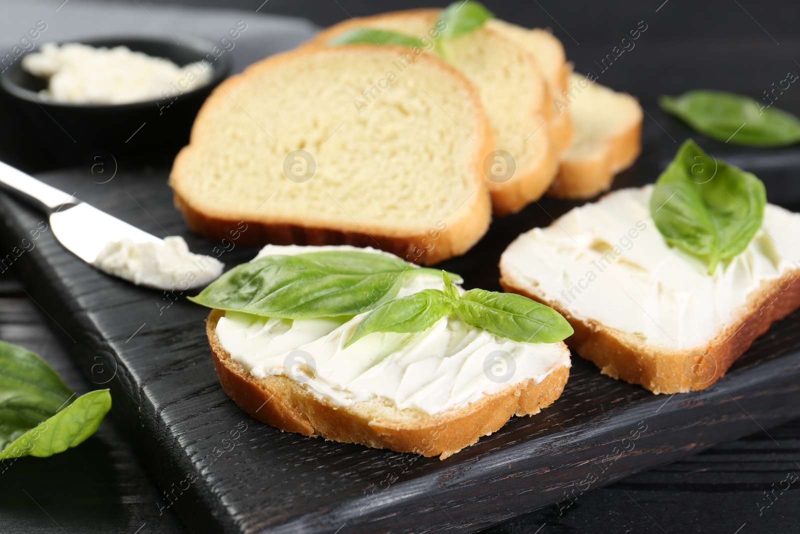 Photo of Pieces of bread with cream cheese and basil leaves on black table, closeup