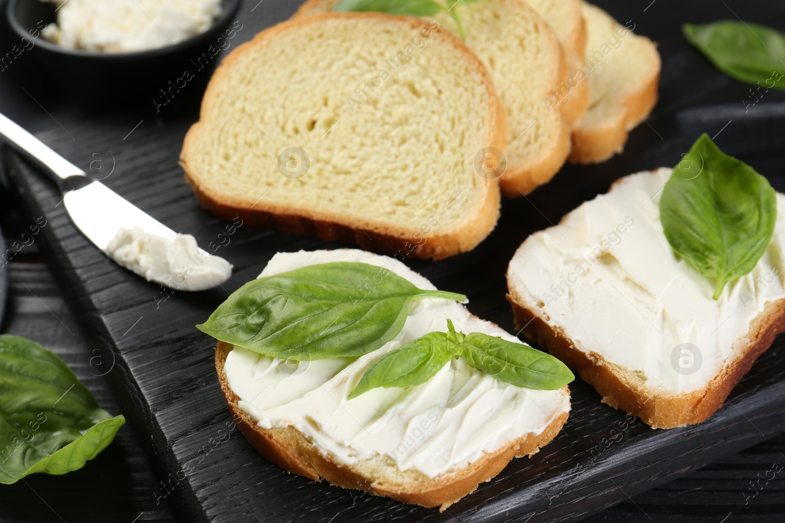 Photo of Pieces of bread with cream cheese and basil leaves on black table, closeup