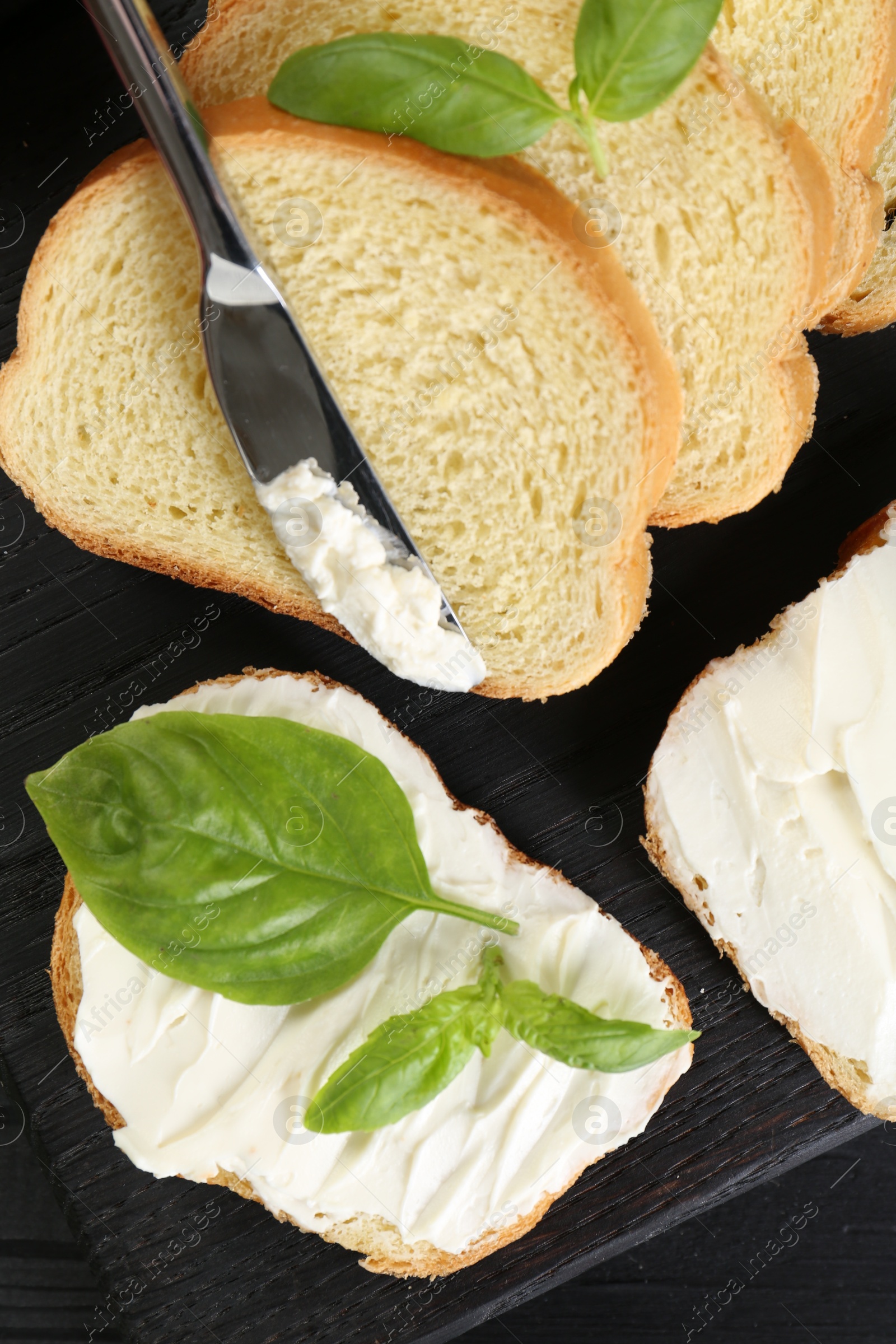 Photo of Pieces of bread with cream cheese, basil and knife on black table, flat lay