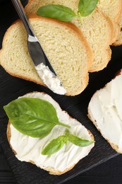 Pieces of bread with cream cheese, basil and knife on black table, flat lay