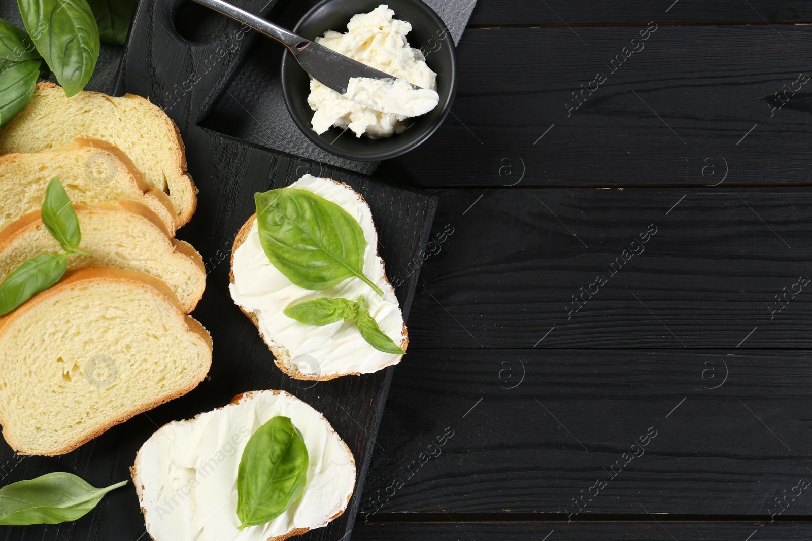 Photo of Pieces of bread with cream cheese and basil served on black wooden table, flat lay. Space for text