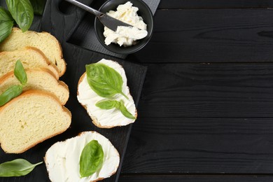 Photo of Pieces of bread with cream cheese and basil served on black wooden table, flat lay. Space for text