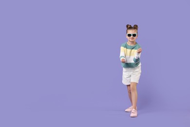 Photo of Cute little girl in sunglasses dancing on violet background, space for text