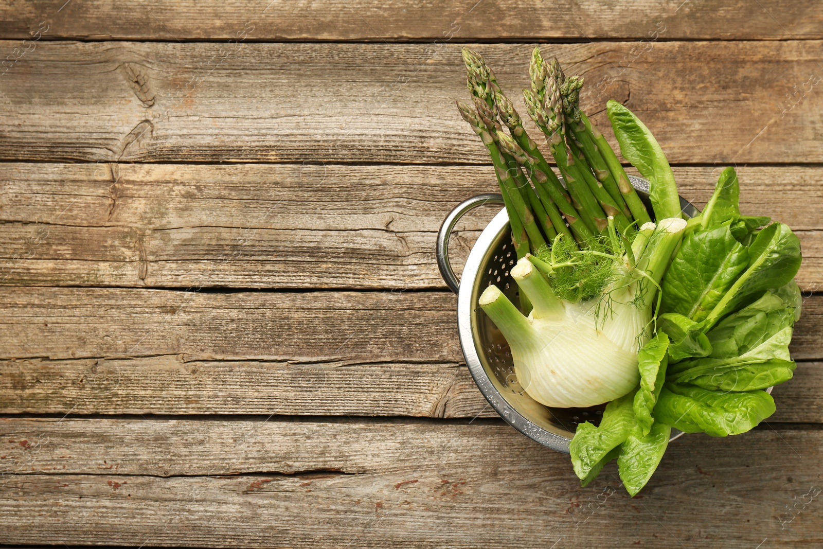 Photo of Metal colander with fennel, lettuce and asparagus on wooden table, top view. Space for text