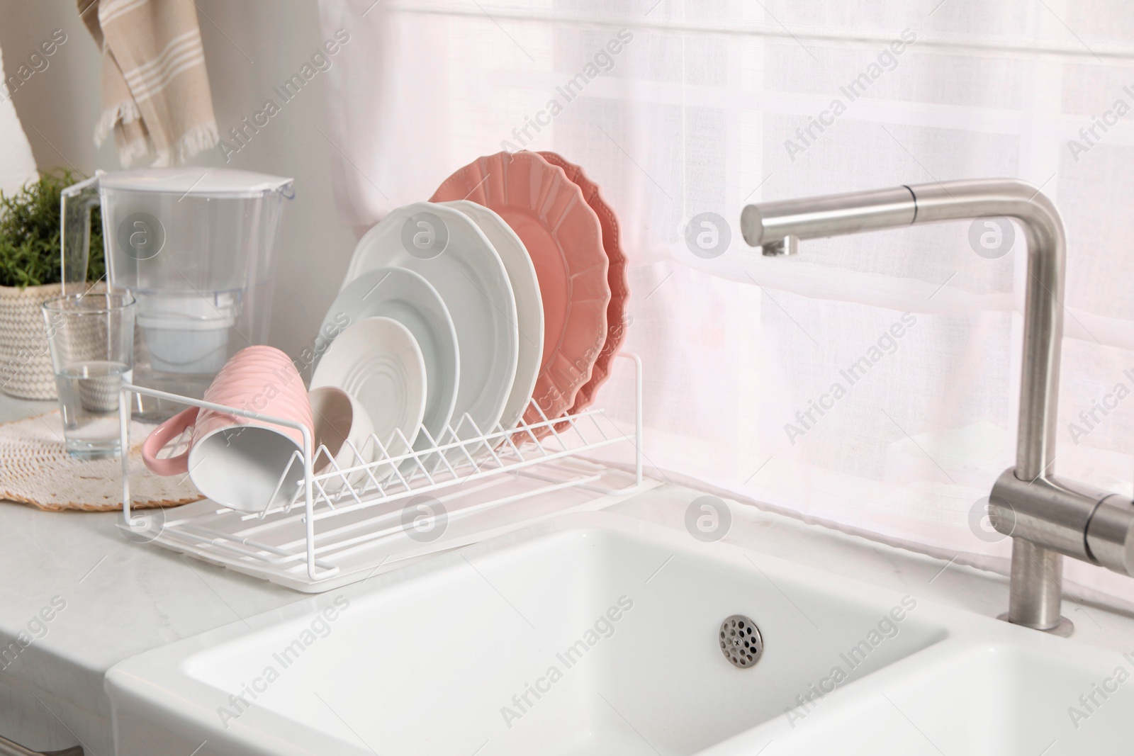 Photo of Drainer with different clean dishware and cup on light table near sink indoors