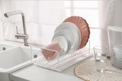 Photo of Drainer with different clean dishware, glass and cup on light table near sink indoors