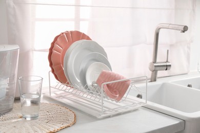 Photo of Drainer with different clean dishware, glass and cup on light table near sink indoors