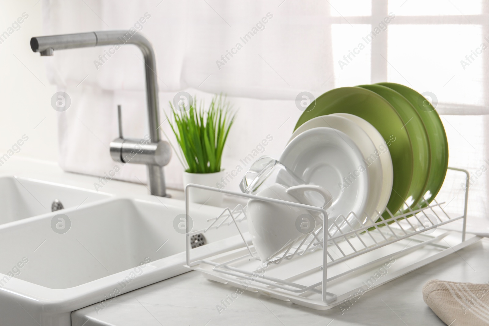 Photo of Drainer with different clean dishware, glass and cup on light table near sink indoors