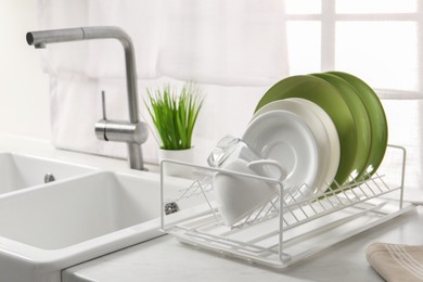 Drainer with different clean dishware, glass and cup on light table near sink indoors