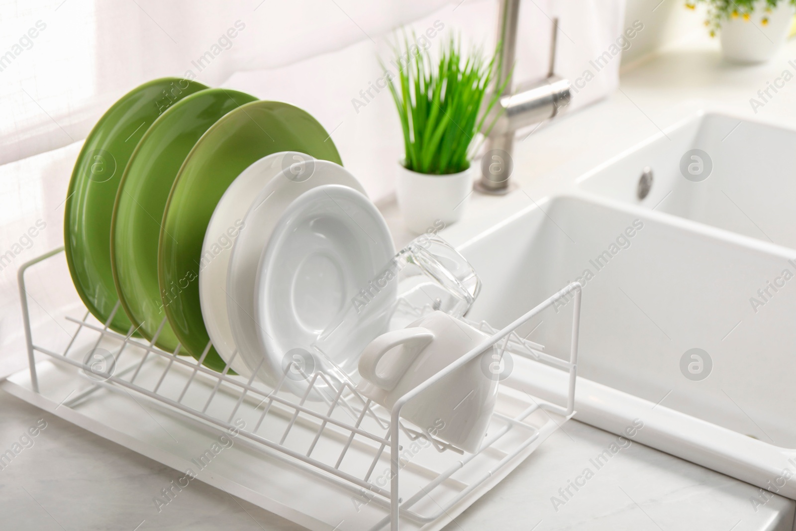 Photo of Drainer with different clean dishware, glass and cup on light table near sink indoors