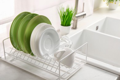 Photo of Drainer with different clean dishware, glass and cup on light table near sink indoors