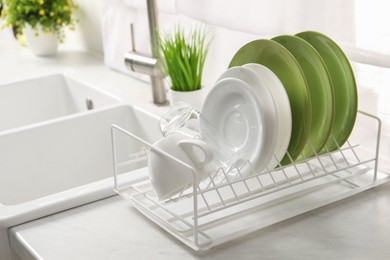 Photo of Drainer with different clean dishware, glass and cup on light table near sink indoors