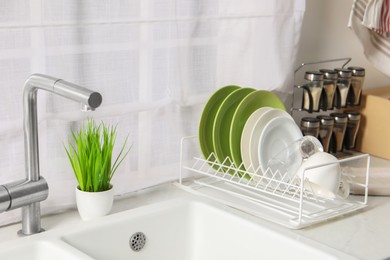 Photo of Drainer with different clean dishware, glass and cup on light table near sink indoors