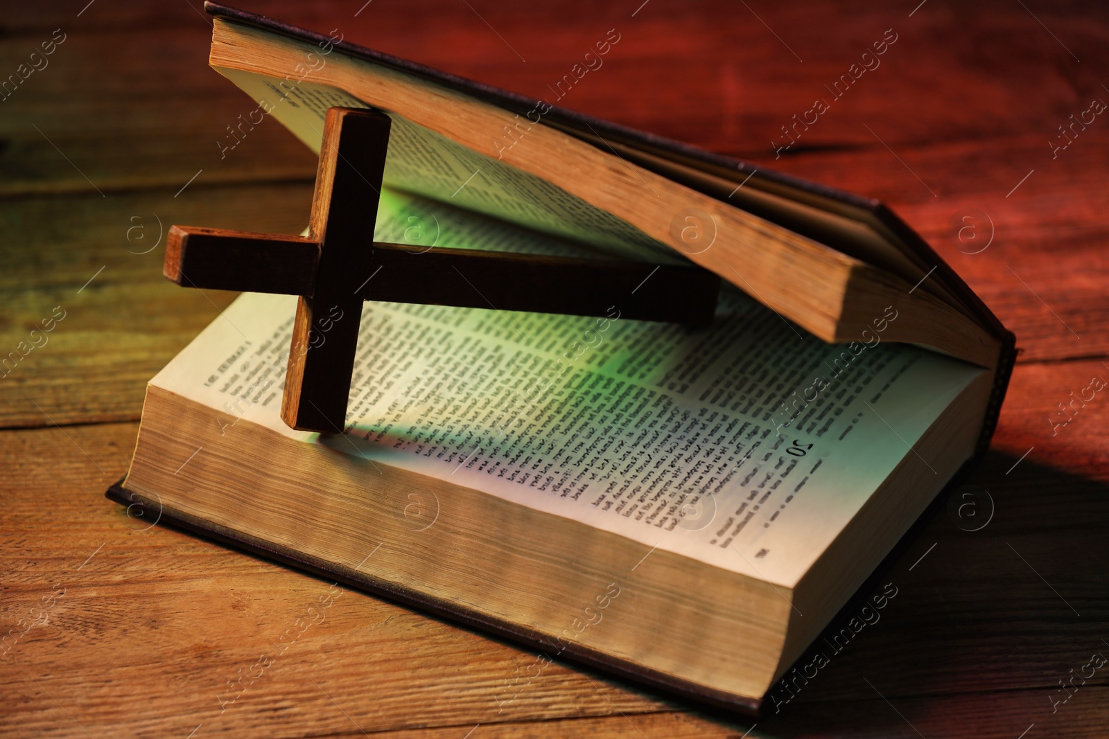 Photo of Cross and Bible on wooden table in color lights, closeup. Religion of Christianity
